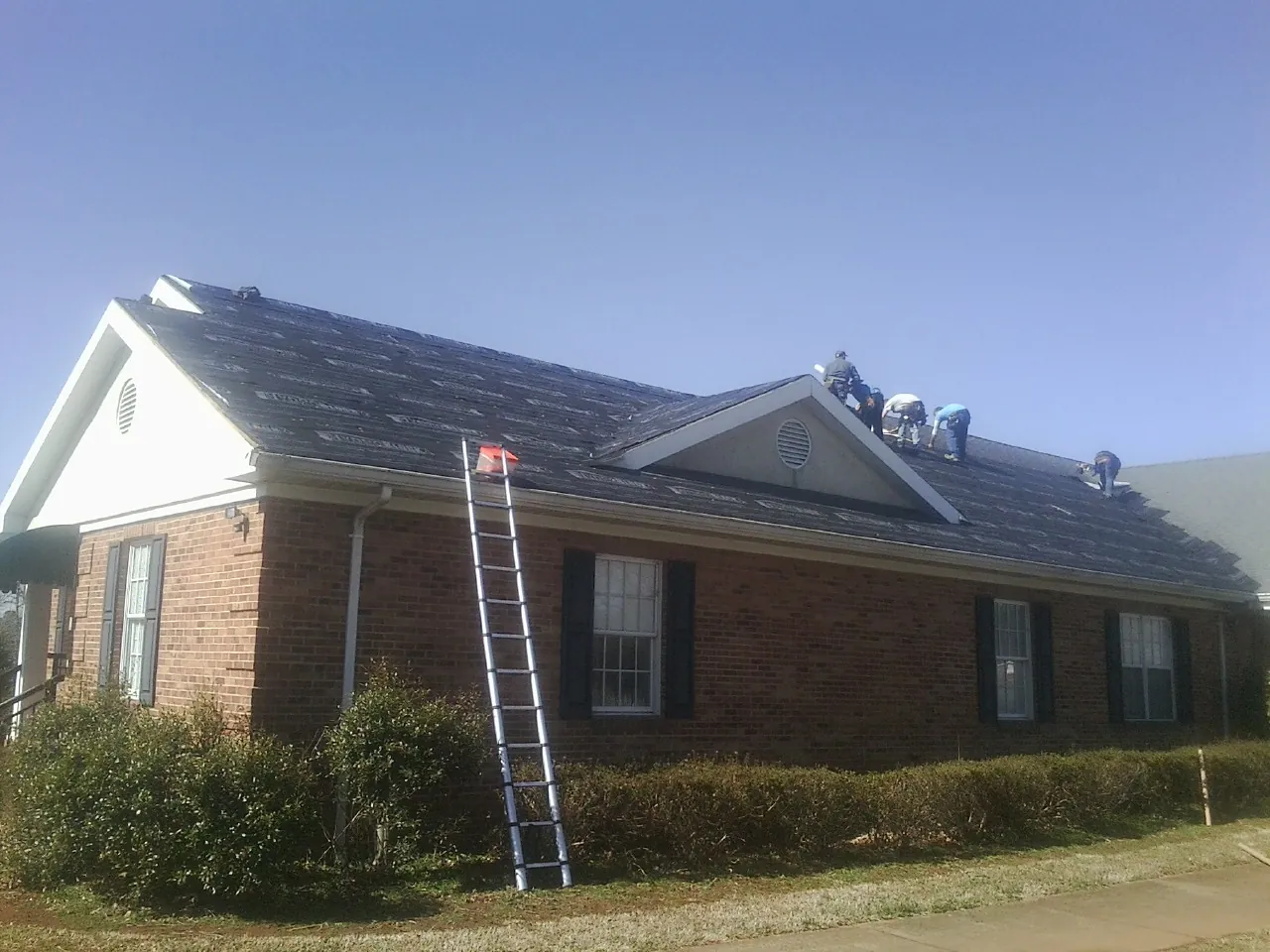 Crew tearing shingles off a brick ranch roof with a ladder set — Emergency Roof Repair work by our crew in Galivants Ferry