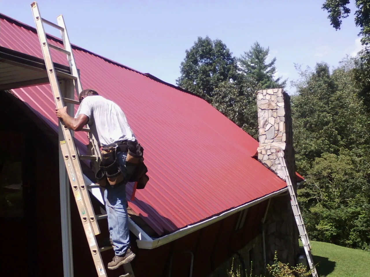 Roofer on a ladder beside a red metal roof and stone chimney — Soffit & Fascia Repair by Roofing Brothers in Galivants Ferry